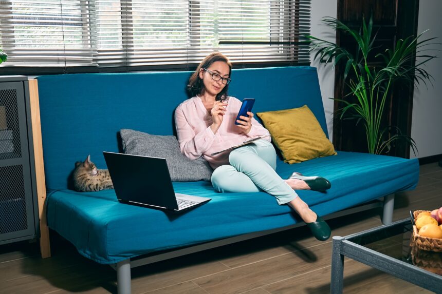 Woman with glasses sitting on sofa with her cat and using smartphone